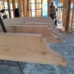 Wooden beams with decorative carvings, organized in a construction site with wooden framing in the background.