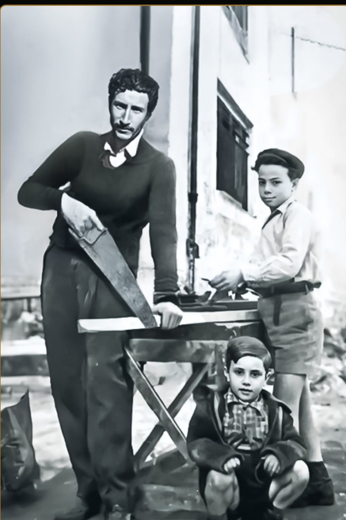 Gerolamo Mazzamuto sawing lumber at an outdoor bench in Sicily, with his son James crouching in front and another boy standing beside the bench